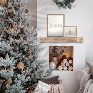 May include: A snowy Christmas tree with brown and black ornaments sits next to a fireplace with a stack of logs. Two framed prints hang above the fireplace, one with a drawing of deer and the other with a simple design.
