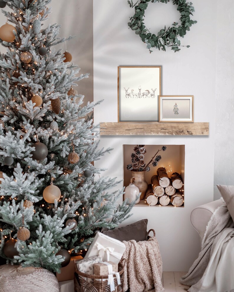 May include: A snowy Christmas tree with brown and black ornaments sits next to a fireplace with a stack of logs. Two framed prints hang above the fireplace, one with a drawing of deer and the other with a simple design.