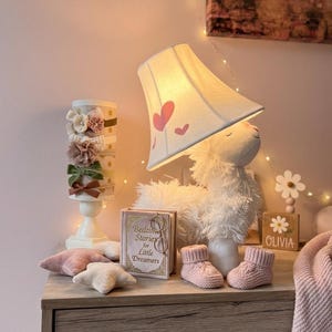 Może przedstawiać: Przytulna scena w pokoju dziecięcym z białą lampą w kształcie lamy z motywem serca na kloszu. Książka zatytułowana "Bedtime Stories for Little Dreamers" leży obok różowych poduszek w kształcie gwiazd i bucików dla niemowląt. Widoczna jest również dekoracyjna lampa z kwiatowymi akcentami.
