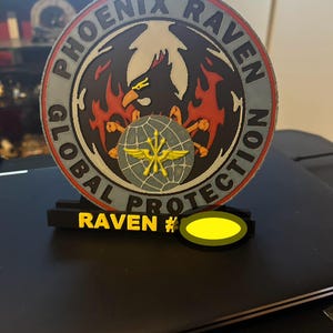 May include: A black and red circular patch with the text "PHOENIX RAVEN GLOBAL PROTECTION" around the edge. A black raven with red flames behind it is in the center of the patch. A smaller patch with a yellow bird and a globe is attached to the larger patch. The text "RAVEN #" is on a black bar below the patch.