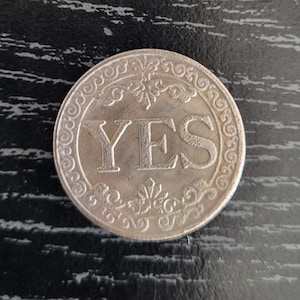 May include: A silver coin with the word "YES" in bold capital letters. The coin has an ornate, decorative border with floral and scroll designs. The coin is circular and appears to be made of metal.