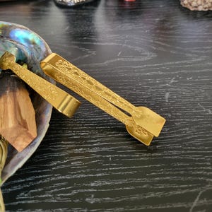 May include: Two gold-colored incense stick tongs with an ornate design. The tongs are lying on a black surface.