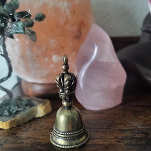 Puede incluir: Una pequeña campana dorada con una parte superior decorativa en forma de corona. La campana está sobre una superficie de madera.