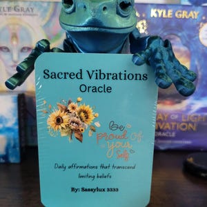 May include: A teal blue card with the text "Sacred Vibrations Oracle" and a floral design featuring sunflowers. The card also has the text "Be proud of your self" and "Daily affirmations that transcend limiting beliefs." The card is held by a green frog figurine.