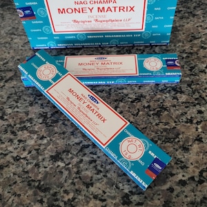 May include: Boxes of Nag Champa Money Matrix incense. The packaging is primarily blue and white, with red accents. The text "Money Matrix" is prominently displayed. The boxes are arranged on a speckled surface.