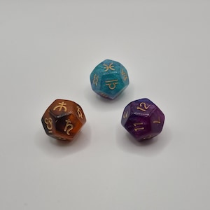Puede incluir: Tres dados poliédricos con símbolos astrológicos y números. Uno es de color naranja-marrón, otro es turquesa y azul, y el tercero es morado. Los dados están sobre una superficie blanca.