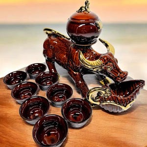 May include: A brown ceramic water buffalo figurine with gold accents, holding a brown ceramic shell. The buffalo has a brown ceramic pot on its back with a gold lid. There are ten brown ceramic cups around the buffalo.