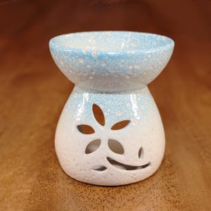 May include: A ceramic essential oil diffuser with a two-tone design. The top is light blue with white speckles, and the base is white with a floral cutout. The diffuser sits on a wooden surface.