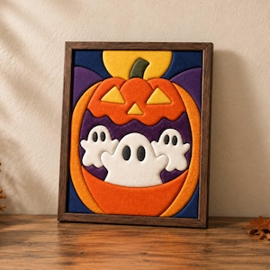 May include: A framed wall art piece featuring a layered felt design of a large orange jack-o'-lantern with a green stem and yellow eyes. Inside the pumpkin, three white ghosts with black eyes are depicted against a deep purple background.
