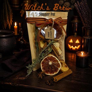 Może przedstawiać: Przezroczysta plastikowa torebka z etykietą Witch's Brew Simmer Pot zawierająca suszone plastry pomarańczy, laski cynamonu i gałązki rozmarynu. Torebka jest przewiązana pomarańczowo-czarną wstążką i posiada małą zawieszkę z napisem Witch's Brew.