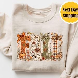 May include: A cream-colored crewneck sweatshirt featuring a large graphic on the front that spells out the word FALL. Each letter is decorated with autumn-themed illustrations including pumpkins, flowers, leaves, mushrooms, and plaid patterns.