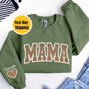 Puede incluir: Una sudadera verde oliva con la palabra "MAMA" en un diseño de estampado de leopardo. La manga tiene el nombre "Amanda" y un corazón con estampado de leopardo. La imagen también incluye una pegatina de "Next Day Shipping".