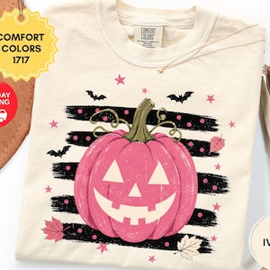 May include: An ivory short-sleeve t-shirt featuring a graphic of a pink jack-o'-lantern pumpkin centered over horizontal black brushstrokes. The design includes small pink stars, black bats, and autumn leaves. The shirt has a Comfort Colors label.