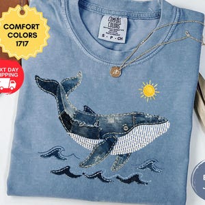May include: A blue short-sleeved t-shirt featuring a graphic of a whale composed of denim-like fabric patches with white striped detailing. The design includes a small yellow sun and dark blue wave accents. A gold-toned necklace rests on the shirt.