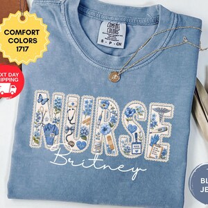 May include: A blue jean Comfort Colors t-shirt with the word "NURSE" in a floral design, with the name "Britney" below. The shirt has a small tag that says "COMFORT COLORS" and "S-P-CH". A gold necklace is draped across the shirt.