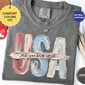 May include: A charcoal gray t-shirt featuring a graphic of the letters USA in a textured, impasto-style paint effect with red, white, and blue colors. A horizontal brushstroke across the letters contains the text 250 years 1776-2026.