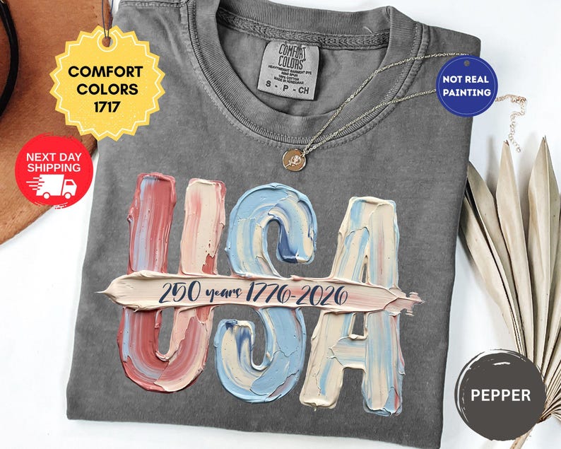 May include: A charcoal gray t-shirt featuring a graphic of the letters USA in a textured, impasto-style paint effect with red, white, and blue colors. A horizontal brushstroke across the letters contains the text 250 years 1776-2026.