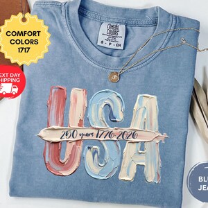 May include: A blue jean colored t-shirt with a Comfort Colors 1717 label. The front features the letters USA in a textured, impasto-style paint effect with red, white, and blue tones. A horizontal brushstroke contains the text 250 years 1776-2026.