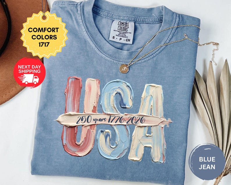 May include: A blue jean colored t-shirt with a Comfort Colors 1717 label. The front features the letters USA in a textured, impasto-style paint effect with red, white, and blue tones. A horizontal brushstroke contains the text 250 years 1776-2026.