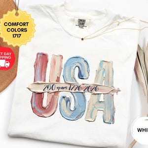 May include: A white short-sleeve t-shirt featuring a graphic of the letters USA in a textured, impasto-style paint effect with red, white, and blue colors. A horizontal brushstroke across the letters contains the text 250 years 1776-2020.