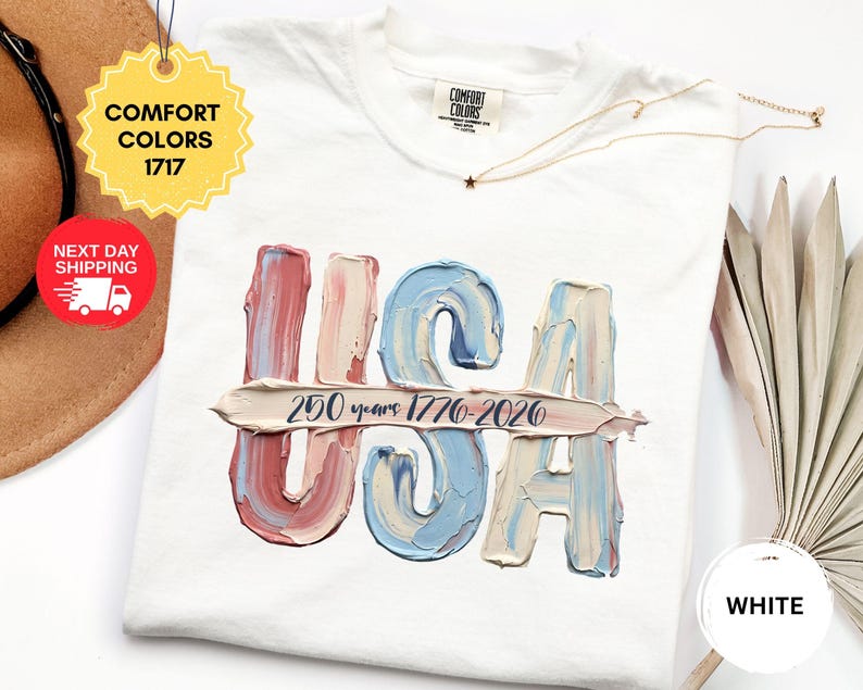 May include: A white short-sleeve t-shirt featuring a graphic of the letters USA in a textured, impasto-style paint effect with red, white, and blue colors. A horizontal brushstroke across the letters contains the text 250 years 1776-2020.