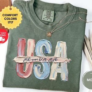 May include: A moss green crew neck t-shirt featuring a graphic of the letters USA in a textured, oil-painted style with red, white, and blue colors. A horizontal brushstroke across the letters displays the text 250 years 1776-2026 in dark blue.
