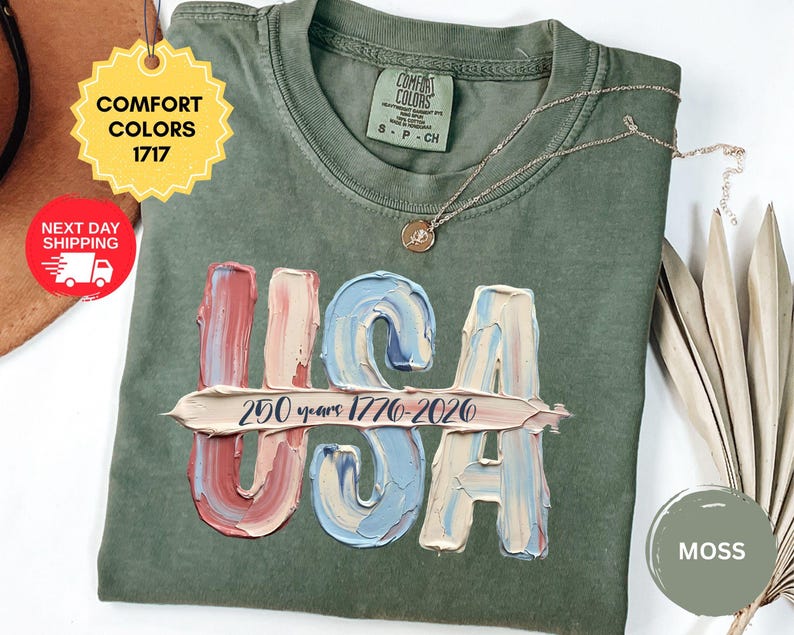 May include: A moss green crew neck t-shirt featuring a graphic of the letters USA in a textured, oil-painted style with red, white, and blue colors. A horizontal brushstroke across the letters displays the text 250 years 1776-2026 in dark blue.