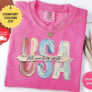May include: A bright pink t-shirt featuring the letters USA in a textured, paint-stroke style with a horizontal brushstroke overlay containing the black text 250 years 1776-2026. The shirt has a Comfort Colors 1717 label and a gold-toned necklace.