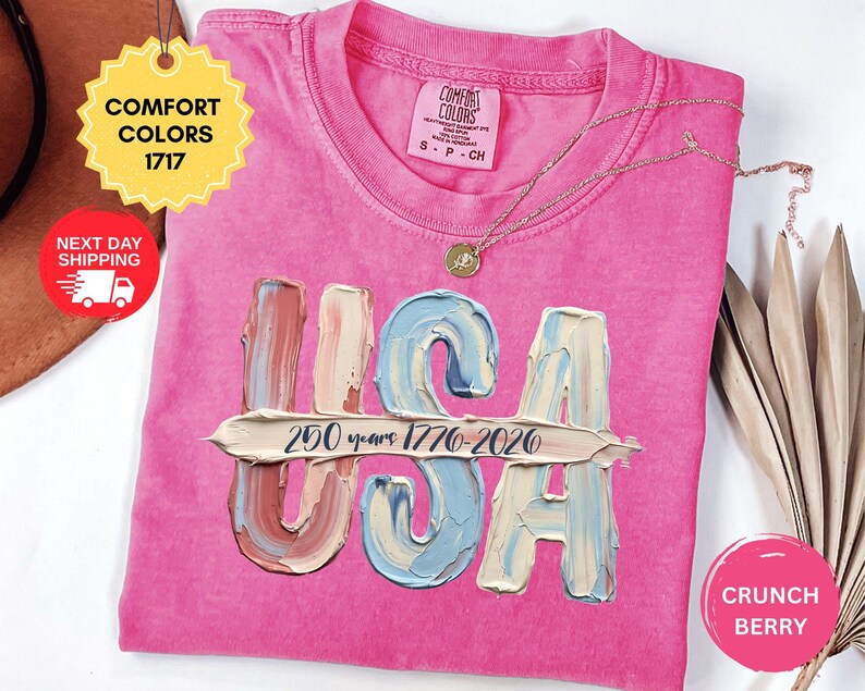 May include: A bright pink t-shirt featuring the letters USA in a textured, paint-stroke style with a horizontal brushstroke overlay containing the black text 250 years 1776-2026. The shirt has a Comfort Colors 1717 label and a gold-toned necklace.