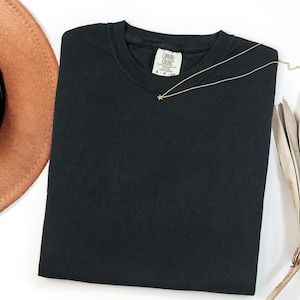 May include: A solid black crew neck t-shirt with a Comfort Colors brand label at the inner neckline. A delicate gold-toned chain necklace featuring a small star-shaped pendant rests on the fabric of the shirt near the collar.