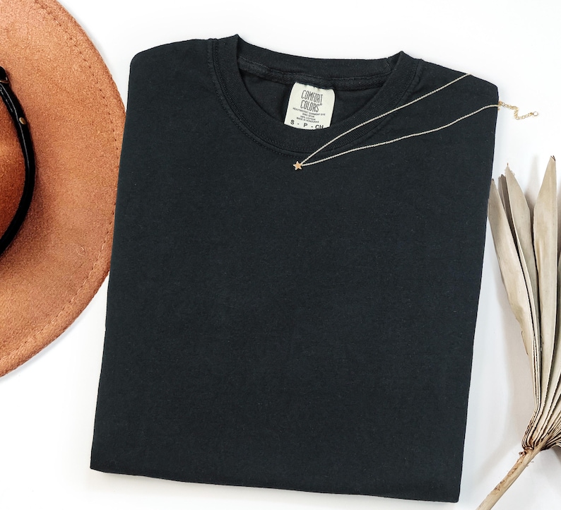 May include: A solid black crew neck t-shirt with a Comfort Colors brand label at the inner neckline. A delicate gold-toned chain necklace featuring a small star-shaped pendant rests on the fabric of the shirt near the collar.