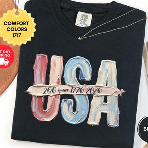 May include: A black t-shirt featuring a graphic of the letters USA rendered in a textured, impasto-style paint effect with red, white, and blue colors. A horizontal brushstroke across the center displays the text 250 years 1776-2026.