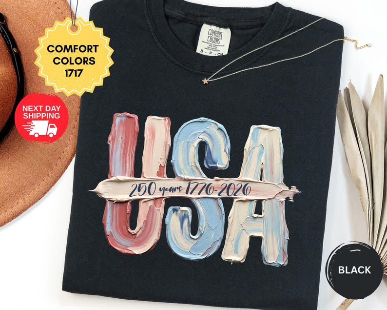 May include: A black t-shirt featuring a graphic of the letters USA rendered in a textured, impasto-style paint effect with red, white, and blue colors. A horizontal brushstroke across the center displays the text 250 years 1776-2026.