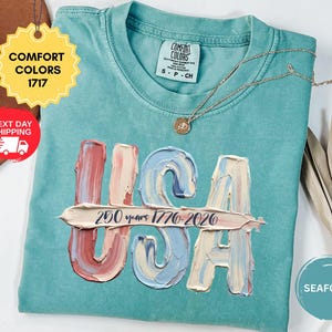 May include: A seafoam green short-sleeve t-shirt featuring a graphic of the letters USA in a textured, impasto-style paint effect with muted red, blue, and cream tones. A horizontal brushstroke across the letters displays the text 250 years 1776-2026.