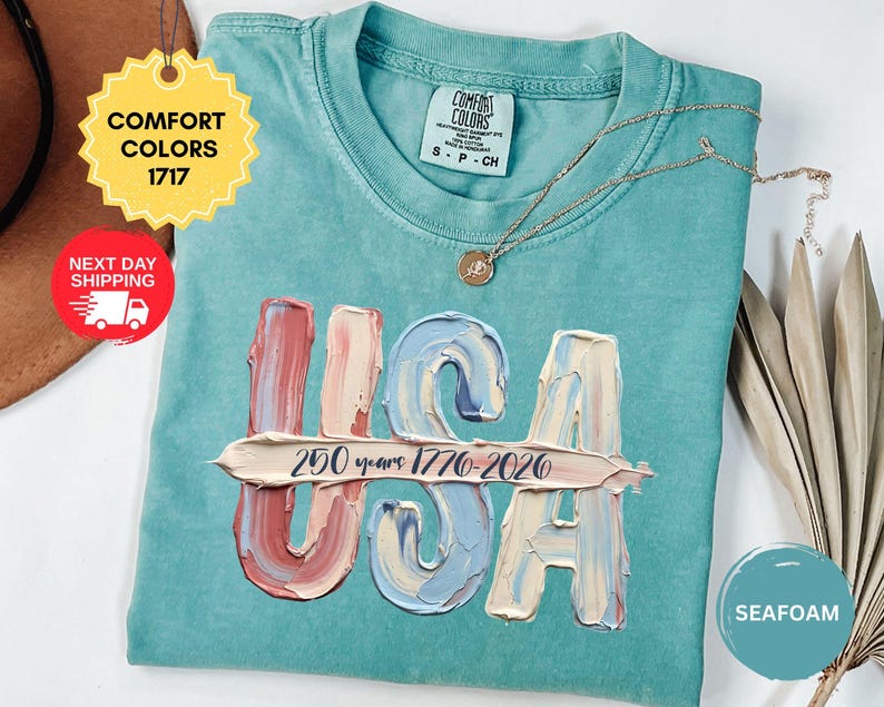 May include: A seafoam green short-sleeve t-shirt featuring a graphic of the letters USA in a textured, impasto-style paint effect with muted red, blue, and cream tones. A horizontal brushstroke across the letters displays the text 250 years 1776-2026.