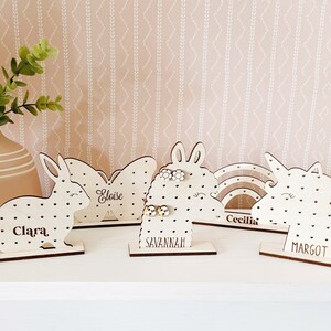 May include: Five wooden earring holders shaped like a bunny, butterfly, unicorn, rainbow, and llama. Each holder has a name on it: Clara, Eloise, Savannah, Cecilia, and Margot. The holders are on a light pink surface with a white pattern.