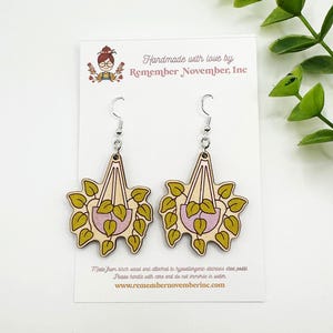 May include: Wooden earrings featuring a plant design with a light pink pot and green leaves. The earrings are attached to hypoallergenic stainless steel posts. The earrings are handmade by Remember November, Inc.