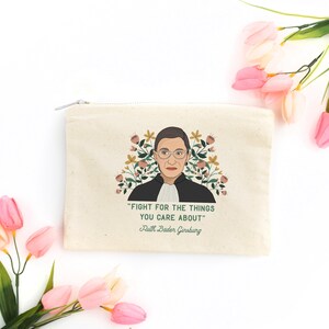 May include: A white canvas zippered pouch with a floral design and an illustration of Ruth Bader Ginsburg. The quote "Fight for the things you care about" is printed below the illustration, along with her name.