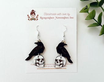 May include: A pair of dangle earrings featuring a black crow perched on a white skull. The earrings are handmade with love by Remember November, Inc.