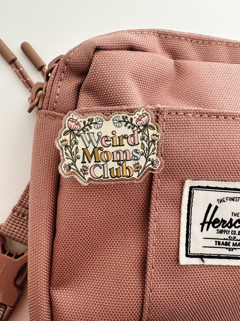May include: A clear acrylic pin with the words "Weird Moms Club" in a whimsical font, surrounded by floral illustrations in pink, yellow, and green. The pin is attached to a dusty rose-colored crossbody bag.