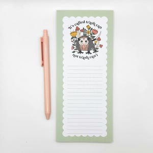 May include: A light green notepad with a scalloped edge, featuring a cartoon opossum illustration with the text "It's called trash can, not trash can't." A matching pink pen rests beside the notepad.