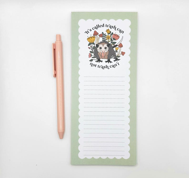 May include: A light green notepad with a scalloped edge, featuring a cartoon opossum illustration with the text "It's called trash can, not trash can't." A matching pink pen rests beside the notepad.