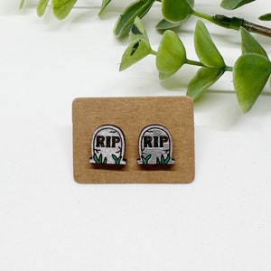 May include: A pair of silver-colored earrings shaped like a tombstone with the text "RIP" on the front. The earrings are on a brown card.