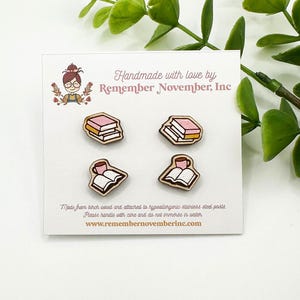 May include: Four wooden pins with book and coffee cup designs. Two pins depict stacks of books with pink covers, and the other two show an open book with a coffee cup. The pins are on a white card with text.