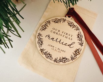 May include: A wooden Christmas ornament with the text "OUR FIRST CHRISTMAS married" inside a floral wreath. The ornament includes the names "JENN + BLAKE" and the date "12.21.23". It rests in a beige pouch with a maroon ribbon.