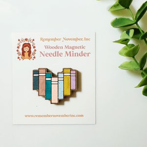 May include: Wooden heart-shaped needle minder with a stack of colorful books design. The needle minder is magnetic and is made by Remember, November, Inc. The text "Remember, November, Inc. Wooden Magnetic Needle Minder" is printed on the card below the needle minder. The website address "www.remembernovemberinc.com" is printed on the bottom of the card.