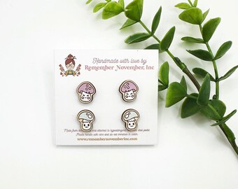 May include: Four wooden mushroom stud earrings on a white card. The mushrooms have pink and blue caps with smiling faces. The card includes the text "Handmade with love by Remember November, Inc."