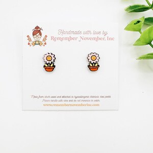 May include: A pair of flower-shaped stud earrings. Each earring features a light pink flower with a yellow center in an orange pot. The earrings are on a white card with text that reads "Handmade with love by Remember November, Inc."