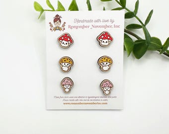 May include: Six wooden mushroom-shaped earrings on a white card. The earrings feature red, yellow, and pink caps with smiling faces. The card reads "Handmade with love by Remember November, Inc."