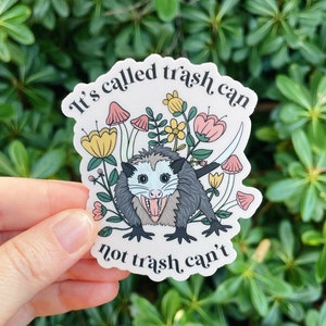 May include: A white sticker with a cartoon possum surrounded by flowers and mushrooms. The text on the sticker reads "It's called trash can not trash, can't".